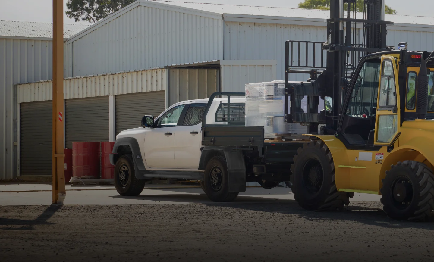 A yellow forklift loads wrapped items onto a white Ford Ranger flatbed pickup truck in an industrial area.