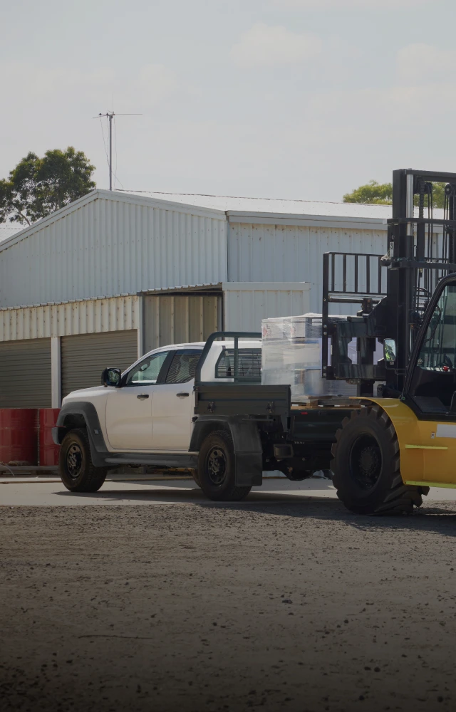 A yellow forklift loads wrapped items onto a white Ford Ranger flatbed pickup truck in an industrial area.