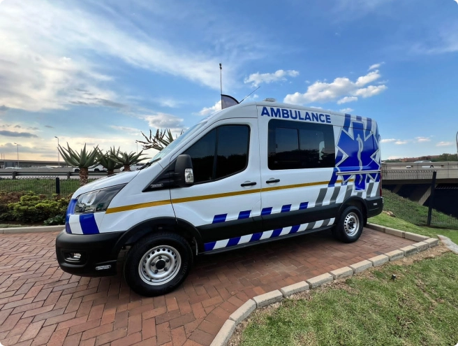 A white ambulance van with blue and white stripes and the Star of Life logo is parked outdoors with a highway overpass in the background.