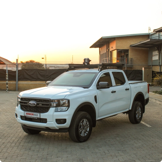 A white Ford Ranger pickup truck with a roof rack, featuring an "ARMORMAX" logo, is parked on a paved surface with a sunset in the background.