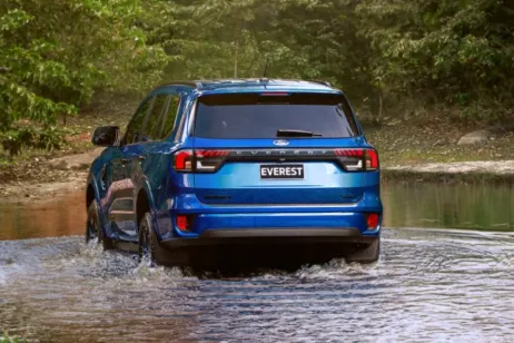 Blue Everest SUV driving through a shallow river surrounded by dense forest 