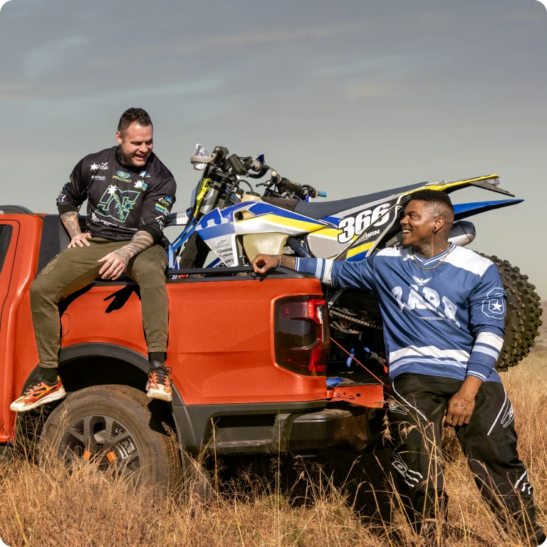 Red pickup truck with dirt bike loaded in rear with two people outdoors.